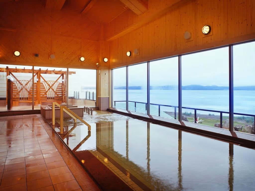 Indoor hot spring bath with large windows overlooking a calm lake and distant mountains.