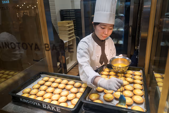 A pastry chef wearing a white uniform and hat glazing rows of custard tarts on baking trays inside a bakery kitchen.