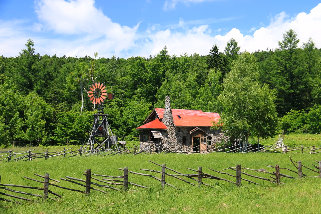 Stone cottage with a red roof and a wooden windmill surrounded by a wooden fence and green trees under a blue sky.