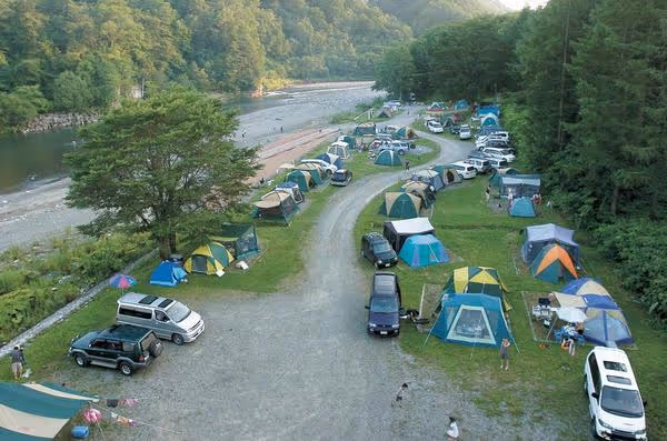 A campground with many tents and cars set along winding paths next to a river and surrounded by forested hills.