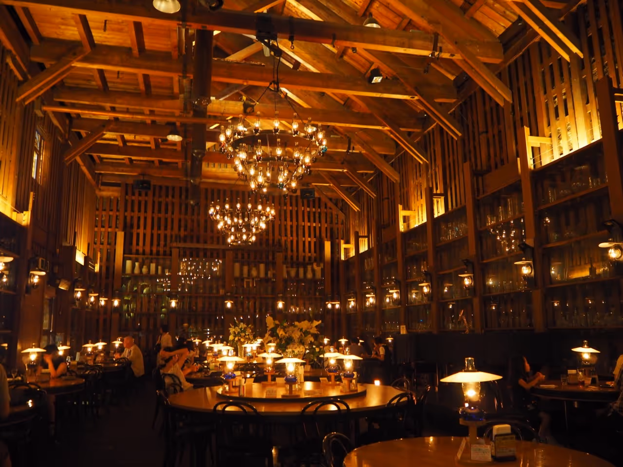 Warmly lit rustic restaurant interior with wooden beams, hanging chandeliers, and tables with vintage-style lamps.