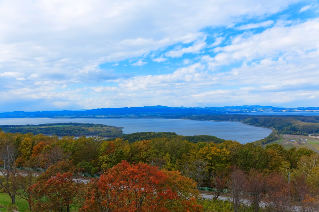 Scenic view of a large lake surrounded by dense forests with autumn-colored trees under a partly cloudy sky.