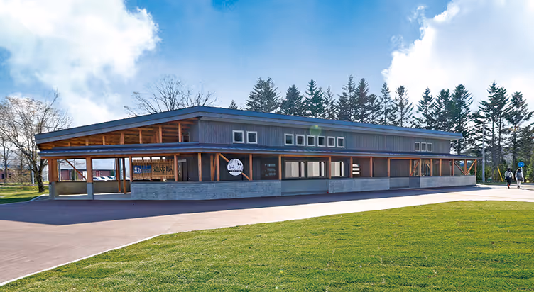Modern single-story wooden building with large windows surrounded by green grass and trees under a blue sky with clouds.