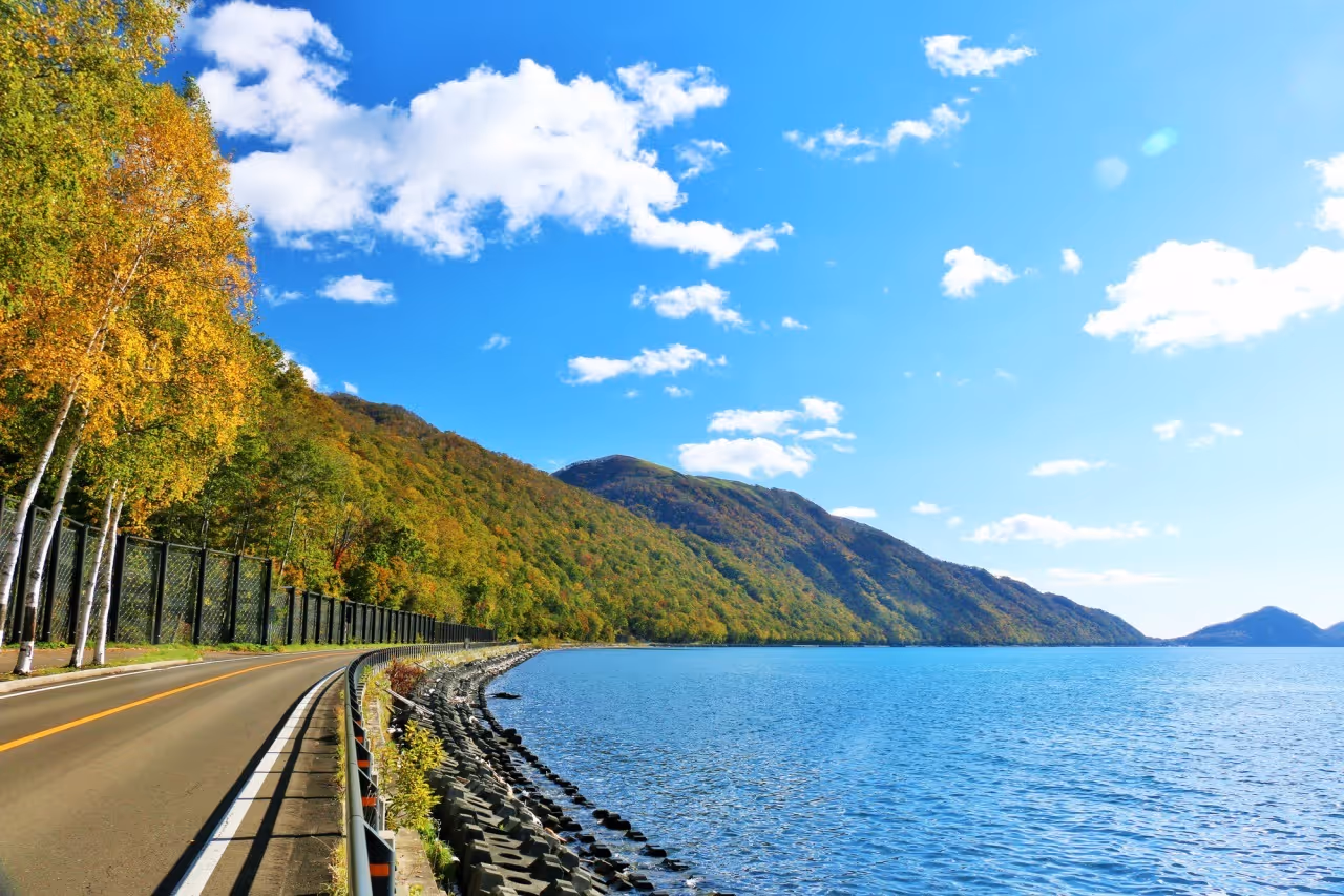 Road bordered by autumn trees curves along the shore of a calm lake with forested hills under a blue sky with scattered white clouds.