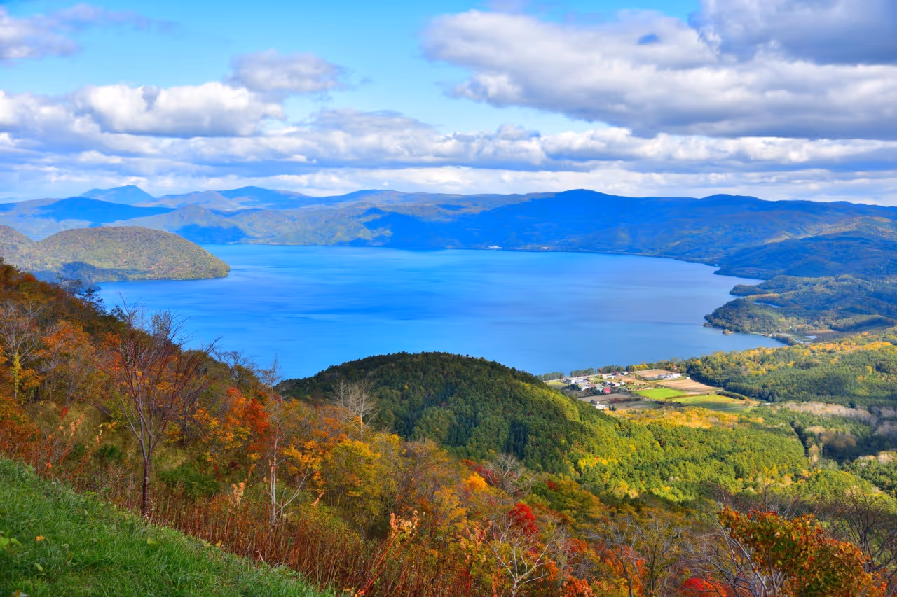 Wide view of a serene blue lake surrounded by autumn-colored forests and distant mountains under a partly cloudy sky.