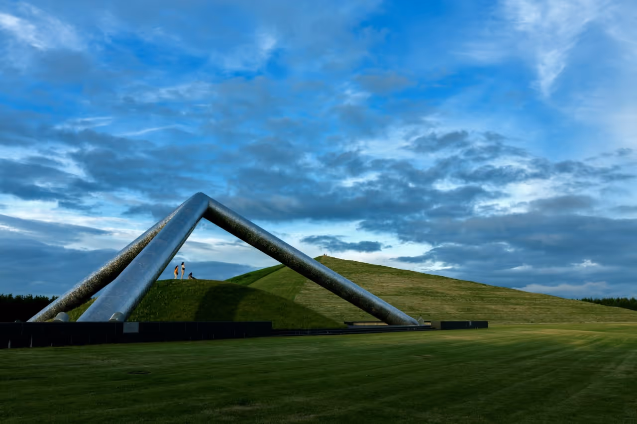 Large outdoor sculpture with two intersecting metallic pipes over grassy mounds under a cloudy blue sky.
