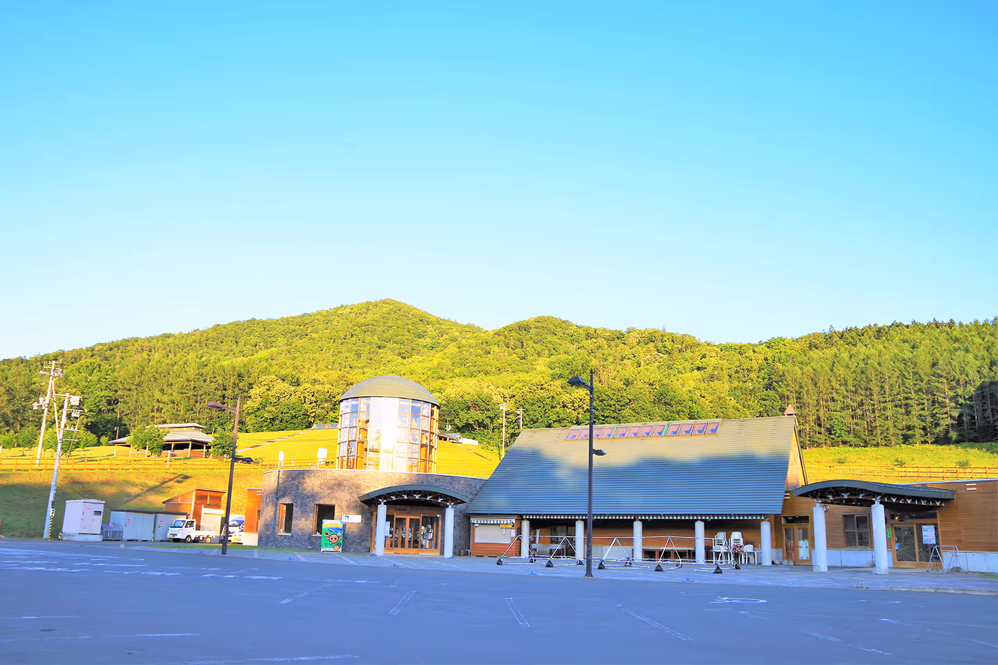 Modern building with a glass cylindrical tower and large sloped roof in front of green forested hills under a clear blue sky.
