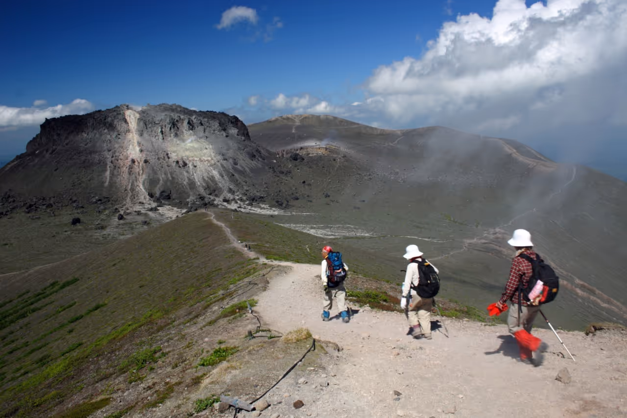 Three hikers walking down a rocky trail on Mount Tarumae with a rugged volcanic mountain and a partly cloudy blue sky in the background.
