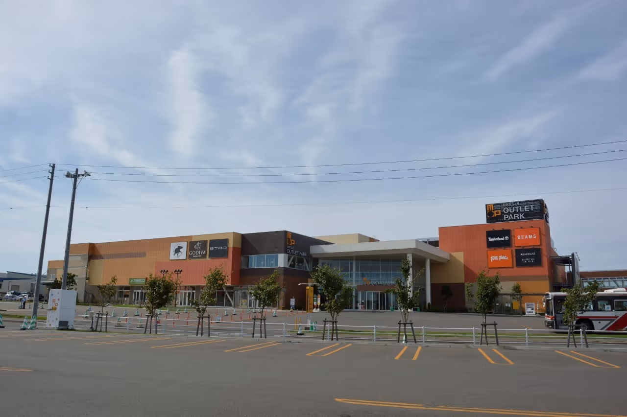 Wide view of Mitsui Outlet Park Sapporo Kitahiroshima building with store signs including Godiva and Timberland, empty parking lot and clear sky.