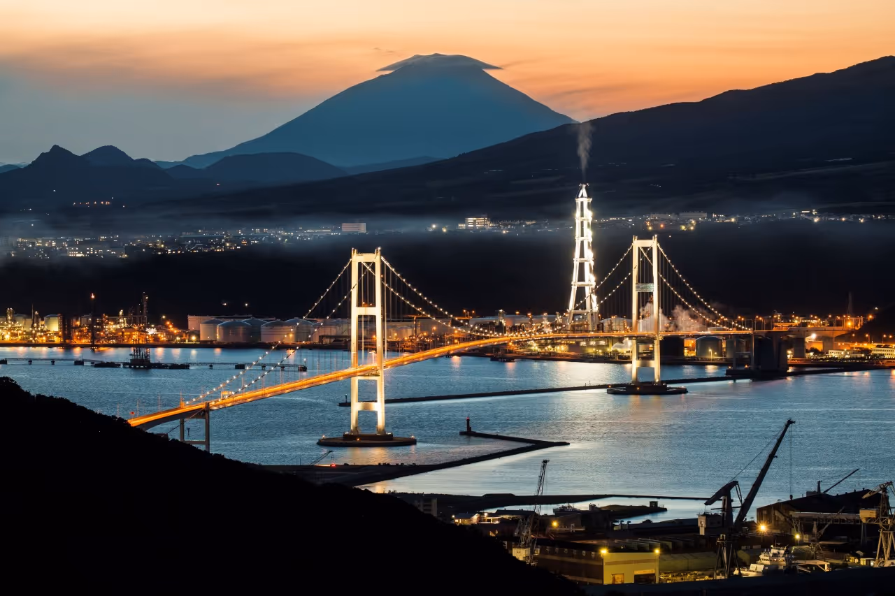 Illuminated suspension bridge over water at dusk with factory and mountain silhouette in the background.