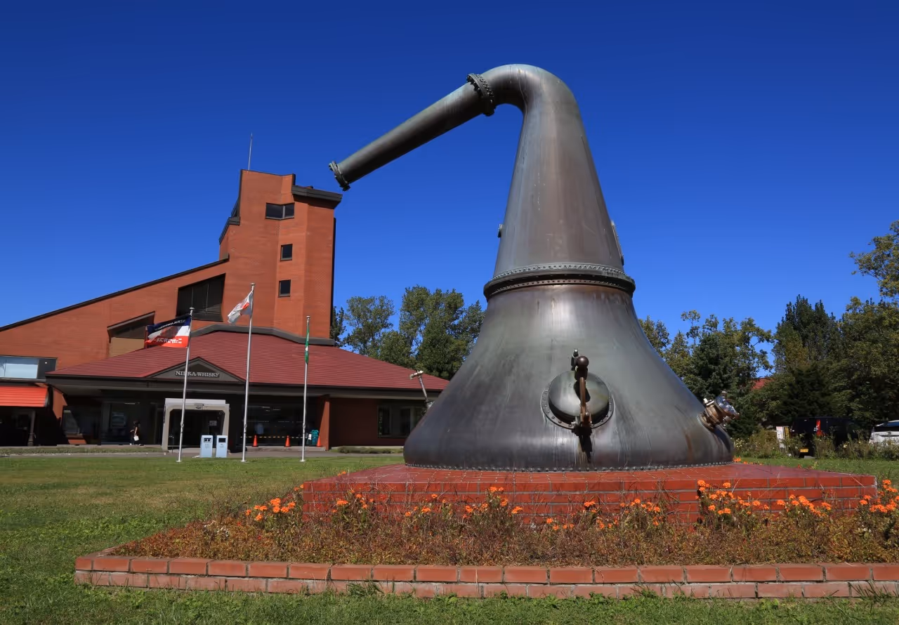 Large copper whisky still displayed outdoors with Nikka Whisky distillery building in the background under clear blue sky.