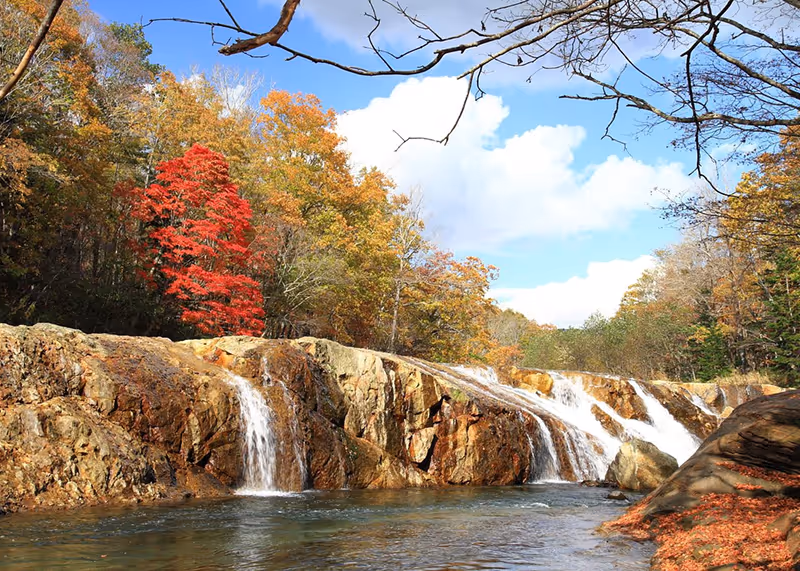 Small waterfalls cascading over rocky ledges surrounded by autumn trees with vibrant red and orange leaves under a blue sky.