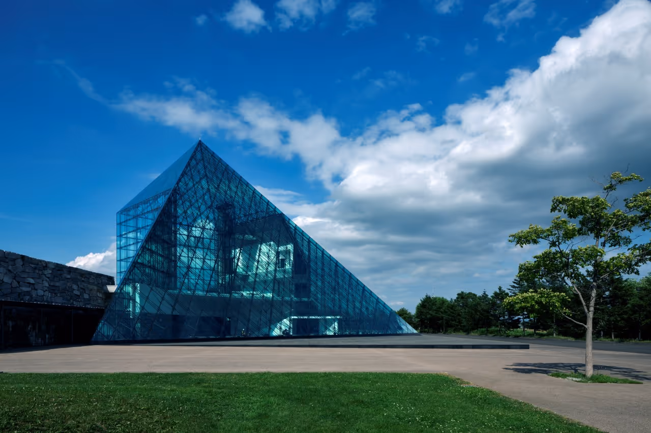 Modern glass pyramid structure with steel framework beside a stone wall under a partly cloudy blue sky.
