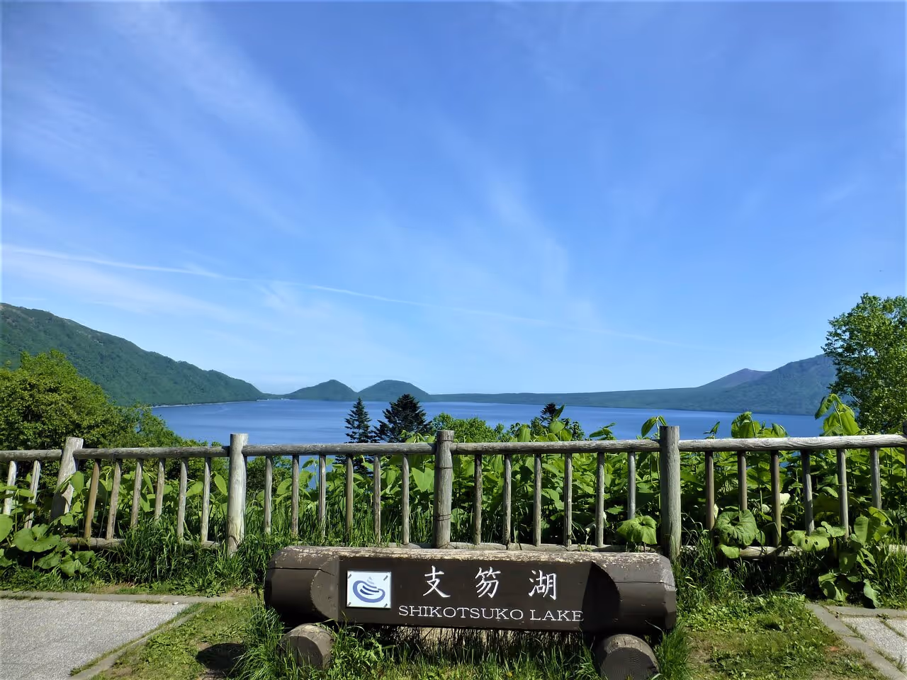 View of Shikotsuko Lake with surrounding green hills and clear blue sky, seen from a wooden fence lookout point.