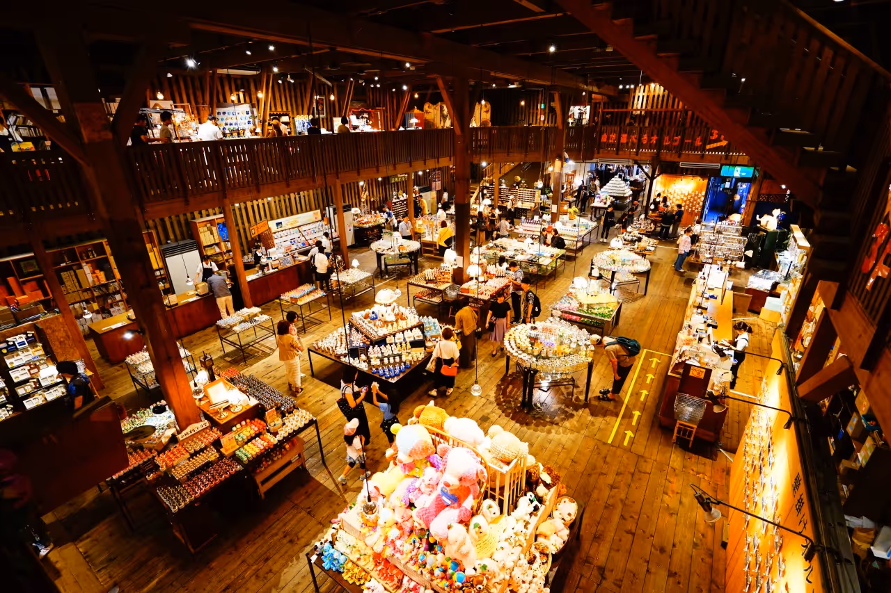 Interior view of a spacious wooden music box museum with multiple display tables featuring colorful music boxes and figurines, and visitors browsing.