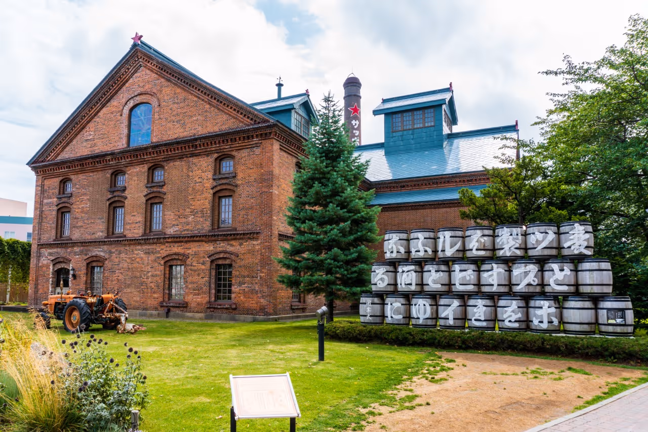 Red brick historic building with a star on its peak, a green tree in front, wooden barrels stacked with Japanese characters, and a vintage orange tractor on the lawn.
