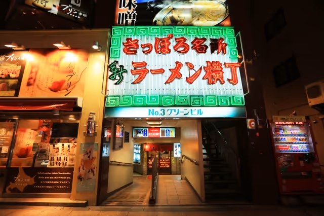 Entrance to Sapporo Ramen Yokocho alley illuminated by neon signs in red and green, with vending machines and stairs visible.