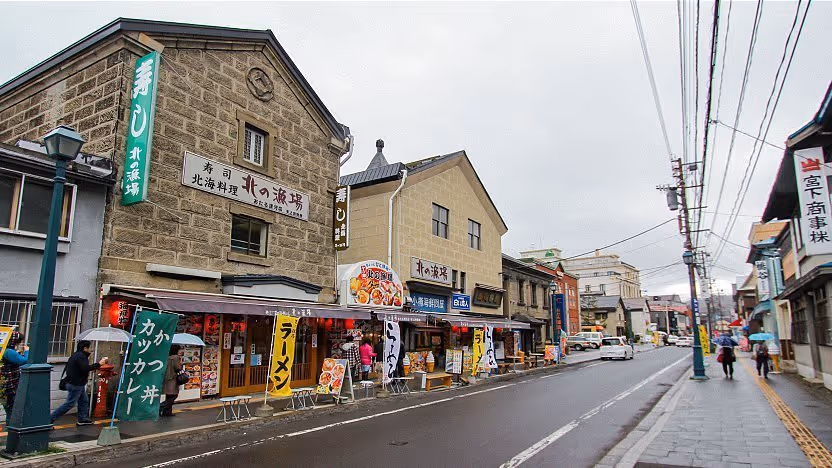 Street view of traditional Japanese buildings with colorful banners advertising food, pedestrians with umbrellas, and a wet road.