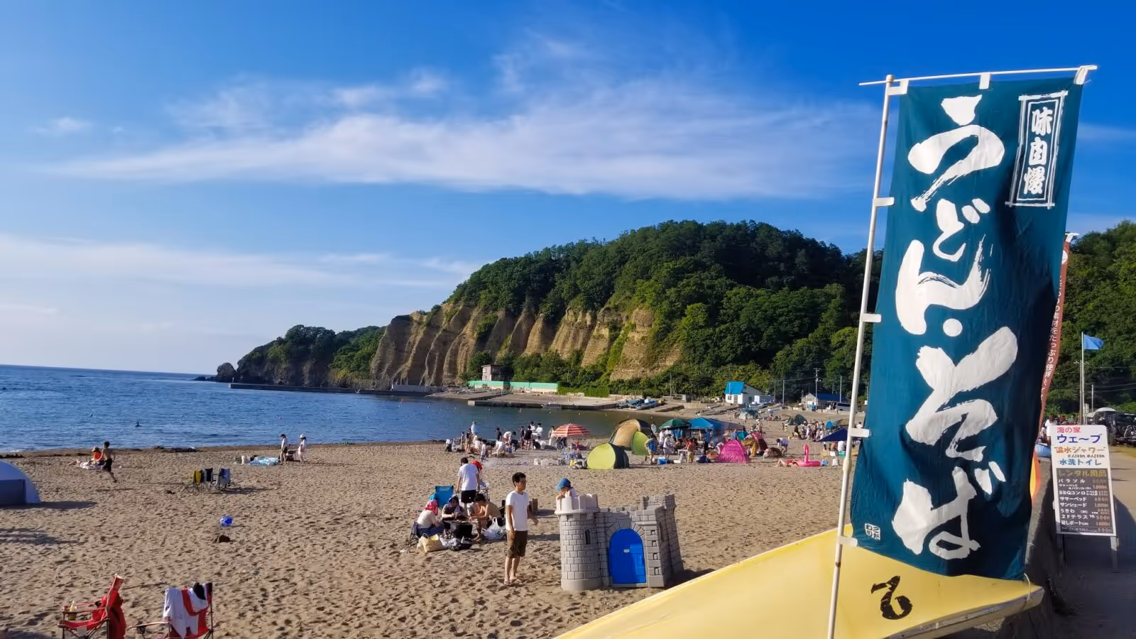 Sandy beach with people relaxing and playing near tents, a blue sky, and green cliffs in the background.