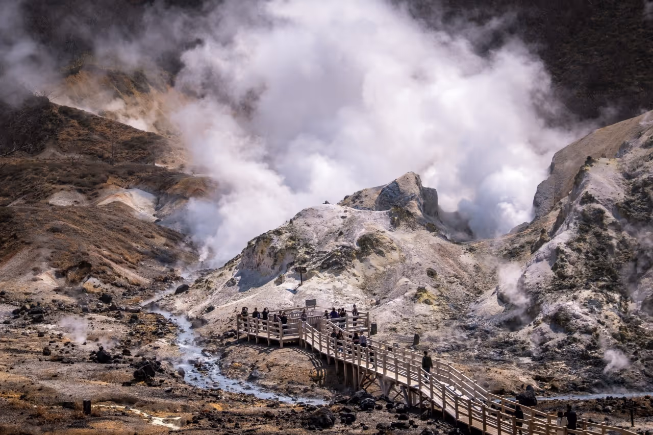 Visitors on a wooden walkway exploring a volcanic area with steaming vents and rocky, barren terrain.