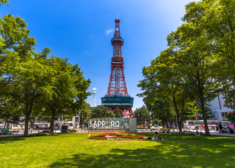 Red and white Sapporo TV Tower standing behind a park with green trees, manicured grass, and flower beds under a clear blue sky.