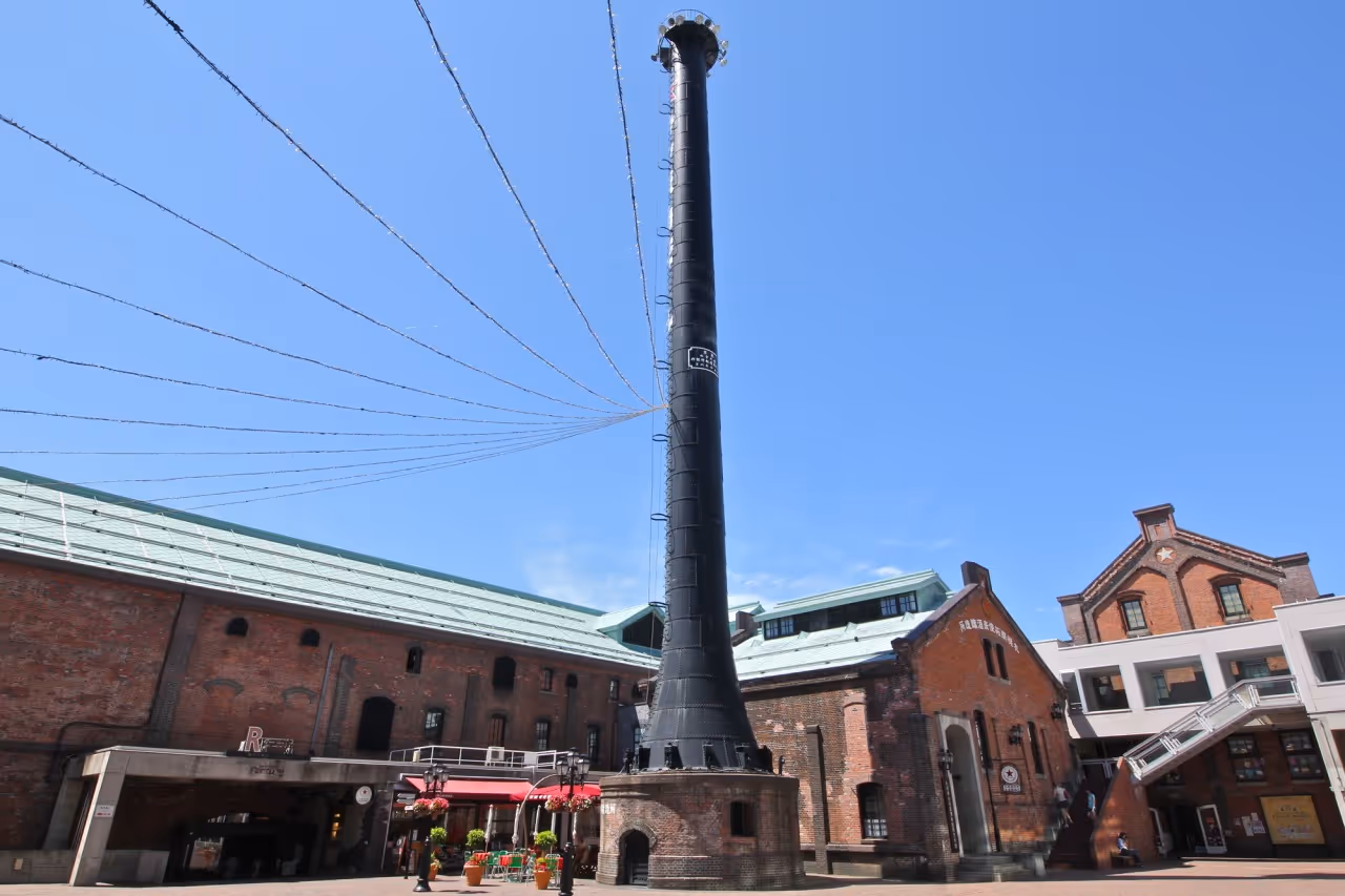 Tall black smokestack surrounded by historic red brick buildings under a clear blue sky at Sapporo Factory.