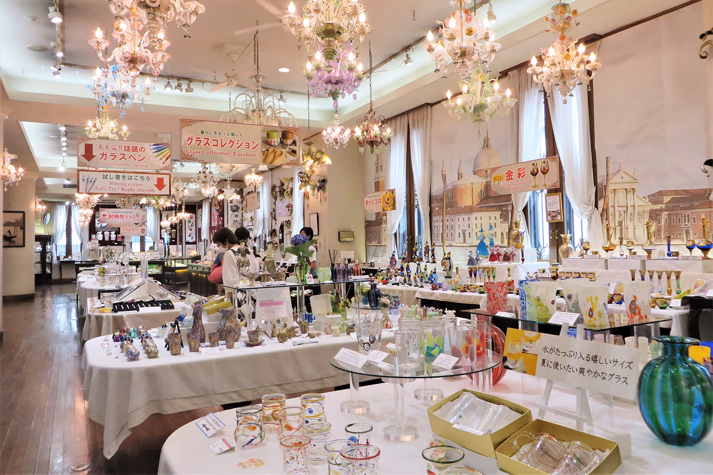 Interior of a brightly lit museum shop displaying colorful Venetian glassware and art pieces on tables with ornate chandeliers overhead.