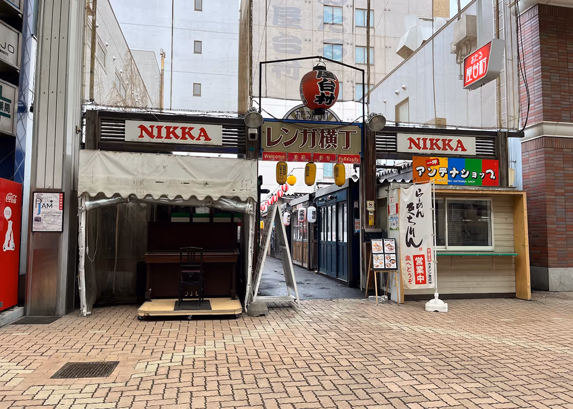 Entrance to a narrow street lined with small shops and lanterns, featuring signs with Japanese characters and the word 'NIKKA' displayed prominently.