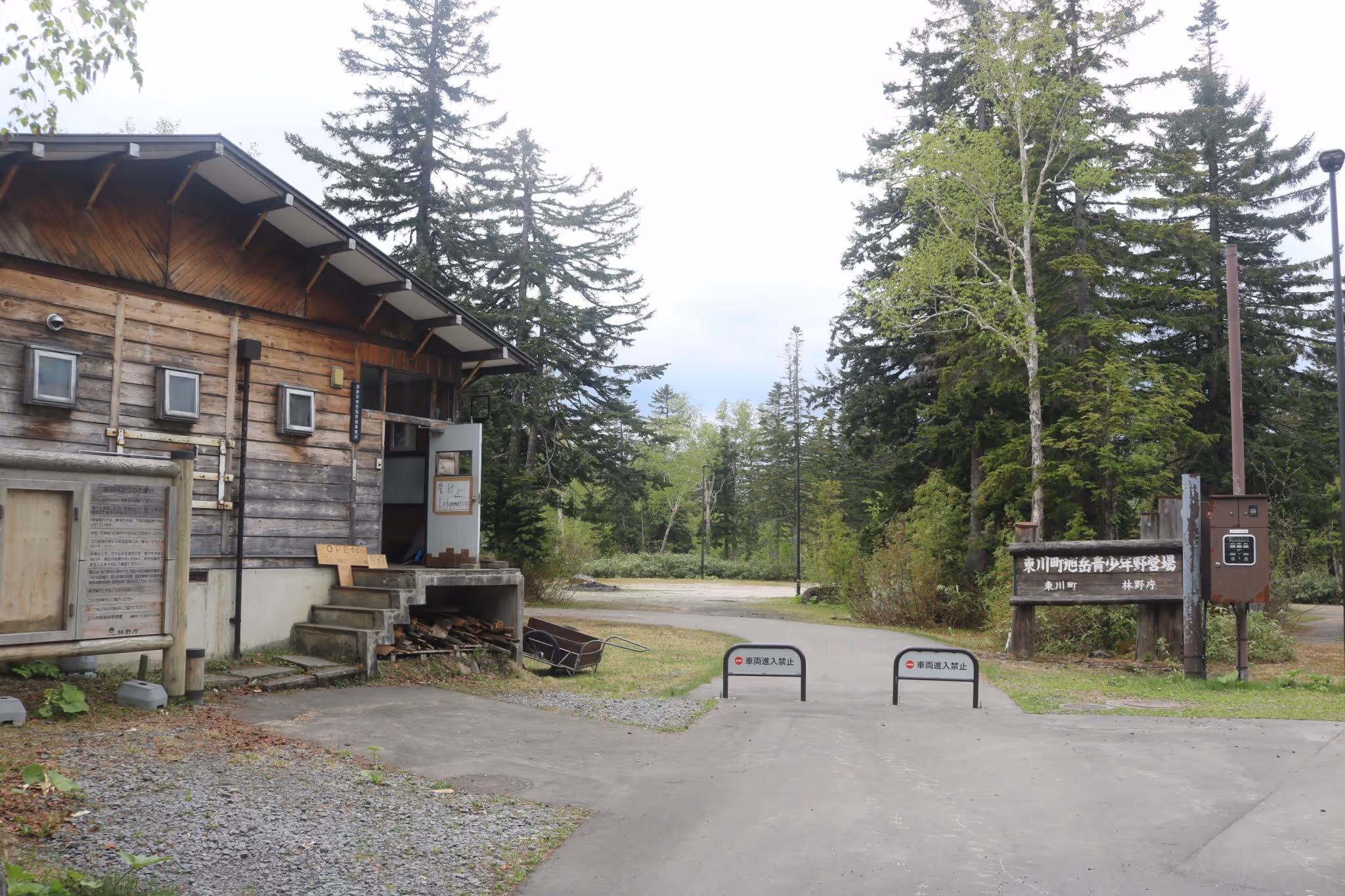Wooden building entrance beside a road blocked by signs, surrounded by green forest trees under a cloudy sky.