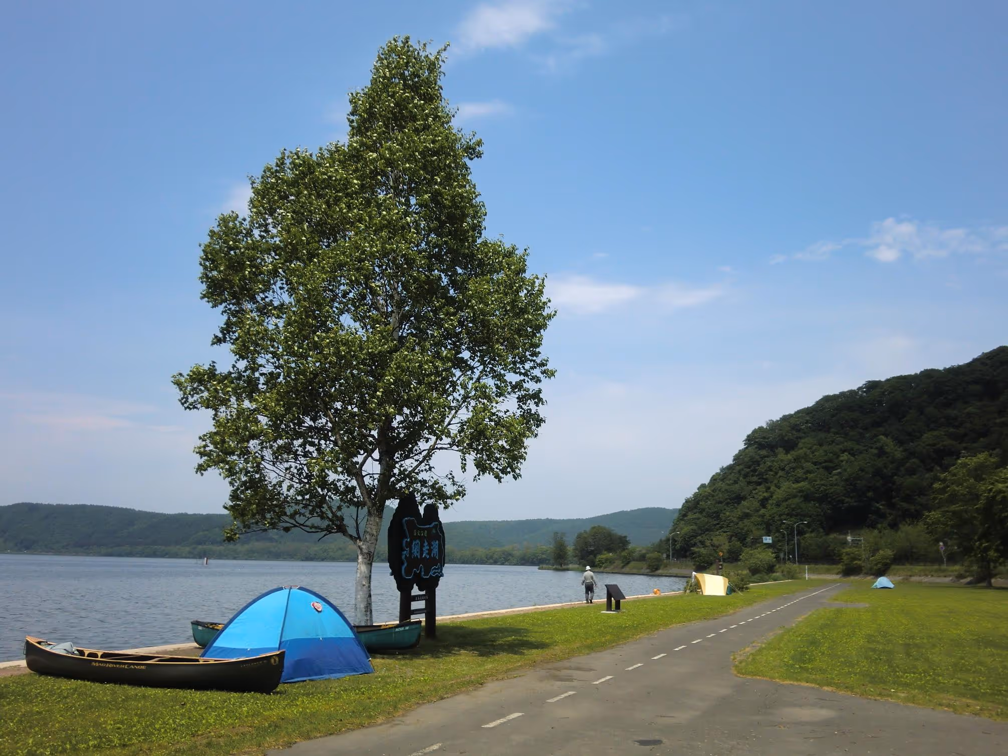 Lakeside campsite with blue tents, canoes on grass, a tree, a lakeshore road, and hills in the background under a clear sky.