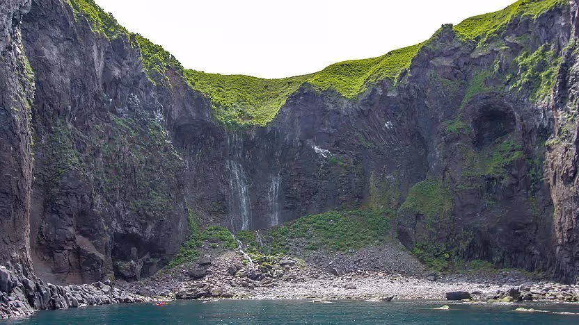 Cliffs covered with green vegetation surrounding a small waterfall flowing into a rocky shore by calm blue water.