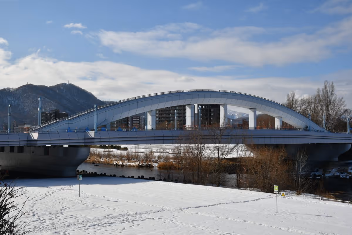 Modern arched bridge spanning a river with snowy ground and mountains in the background under a partly cloudy sky.