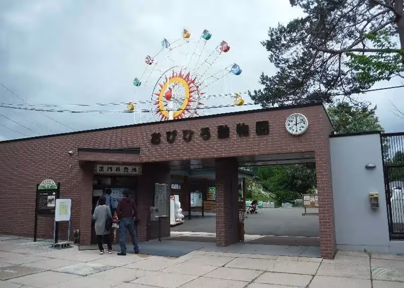 Entrance gate to a zoo with a clock and a colorful Ferris wheel visible in the background under a cloudy sky.