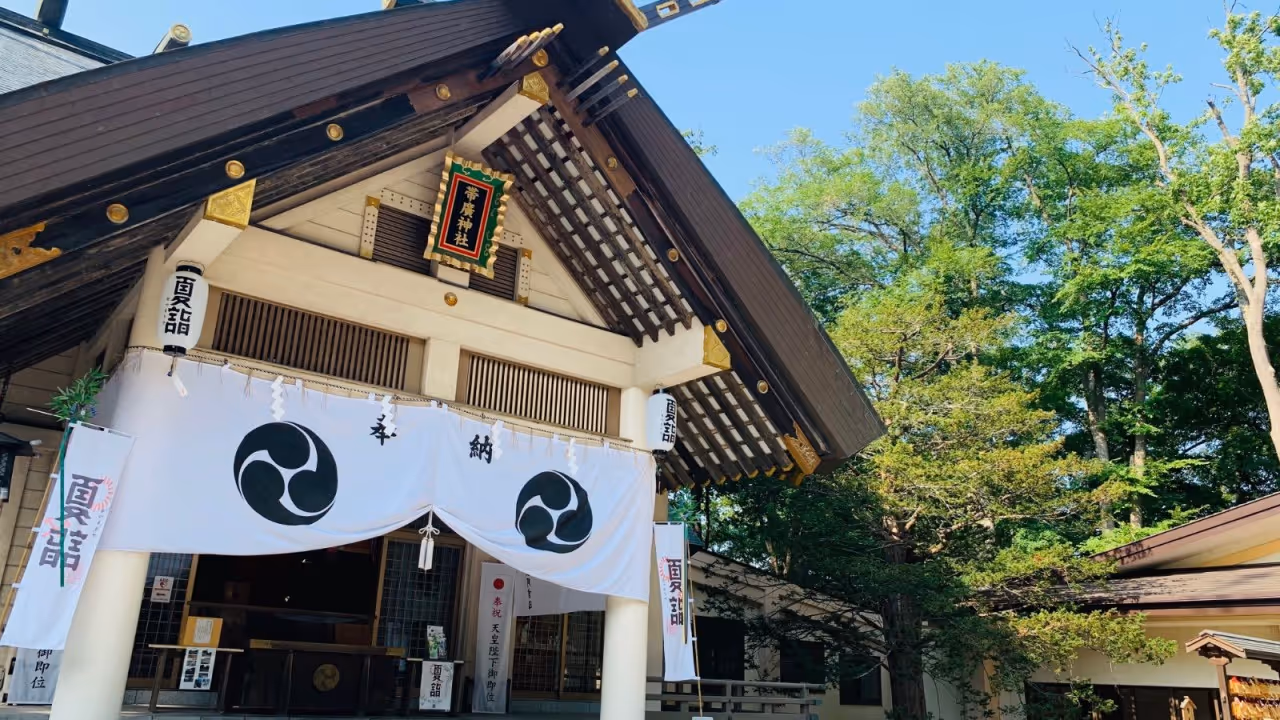 Entrance of a Japanese shrine with white banners displaying black circular symbols under a dark wooden roof, surrounded by green trees.
