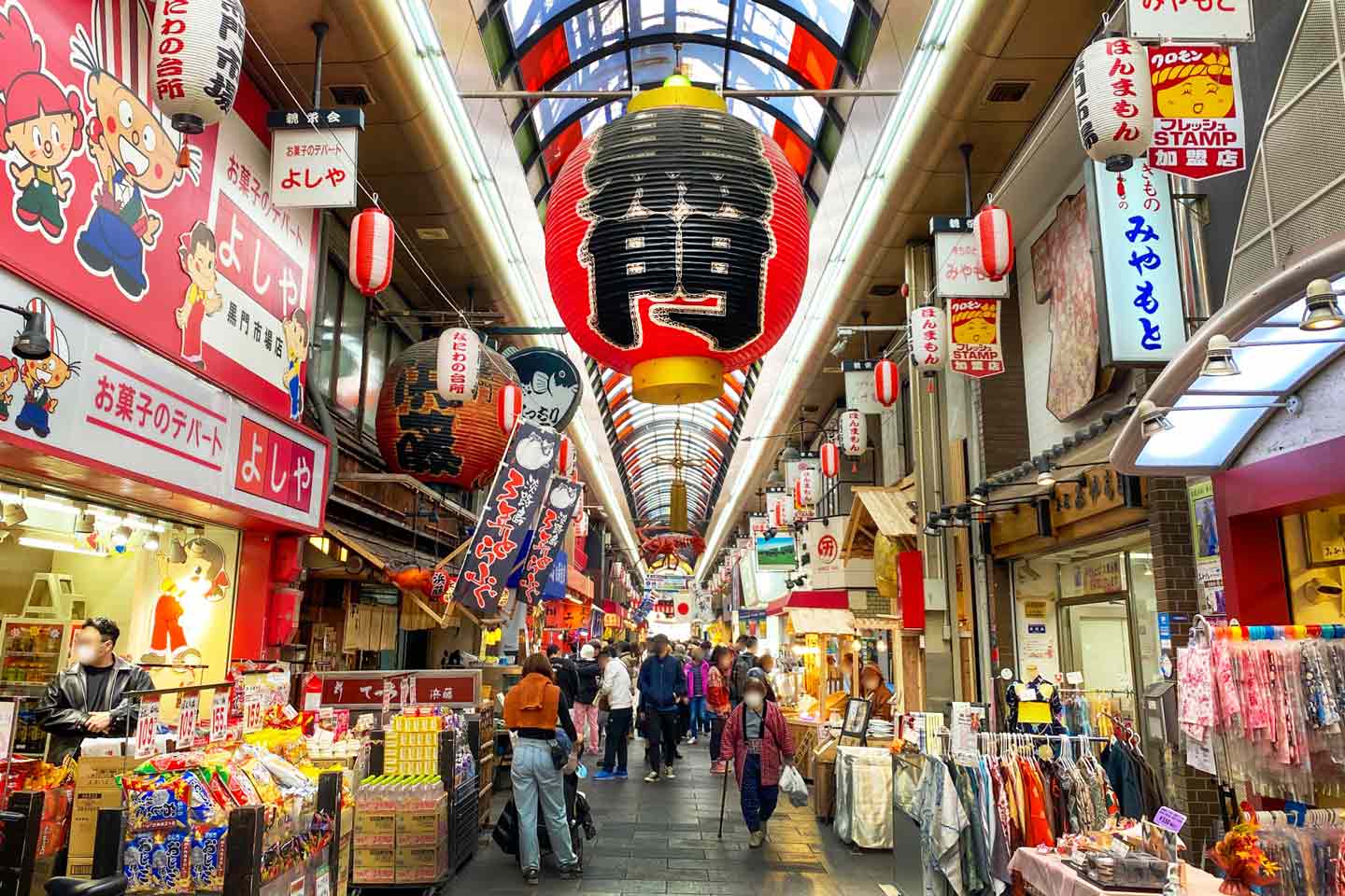 Busy covered shopping street in Japan with hanging red and black lanterns, colorful shop signs, and people walking.