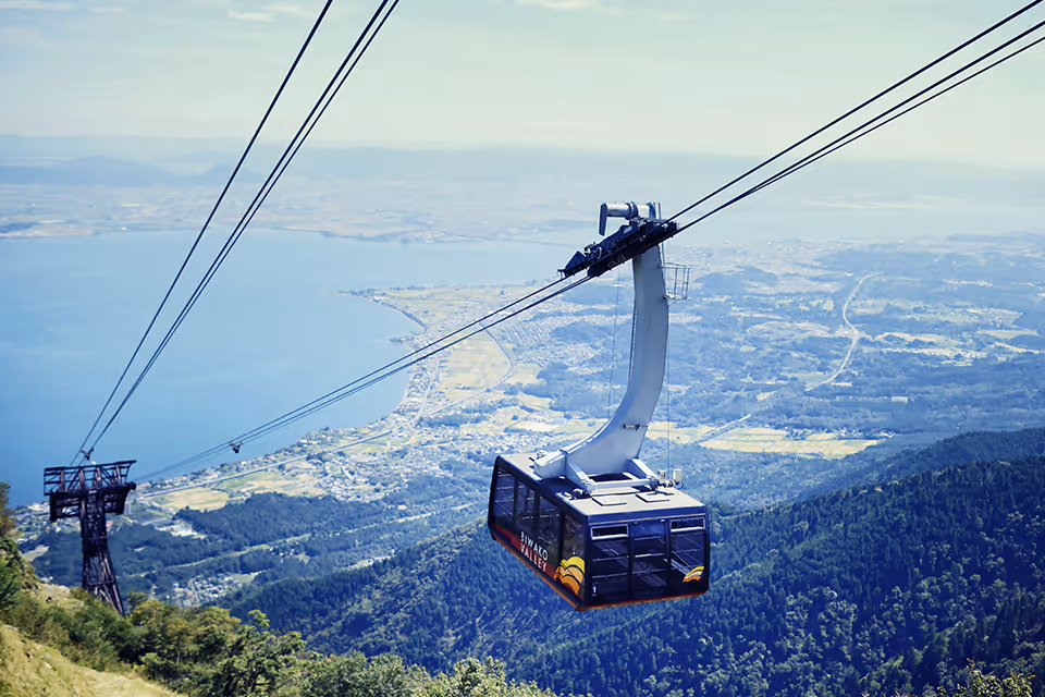 Cable car labeled 'Biwako Valley' suspended over forested mountains with a lake and distant landscape in the background.