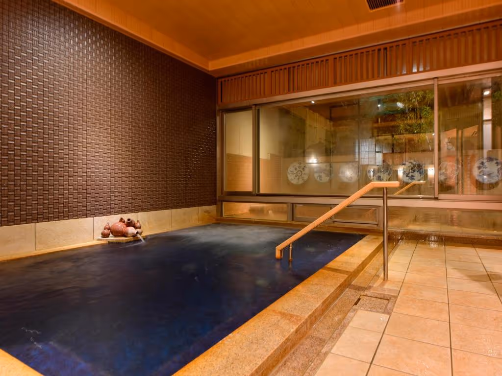 Indoor Japanese onsen bath with dark water, tiled floor, wooden ceiling, and decorative water spout and window displaying ceramic plates.
