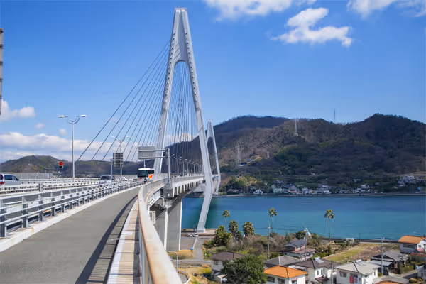 Cable-stayed bridge over blue water leading towards forested hills under a partly cloudy sky.