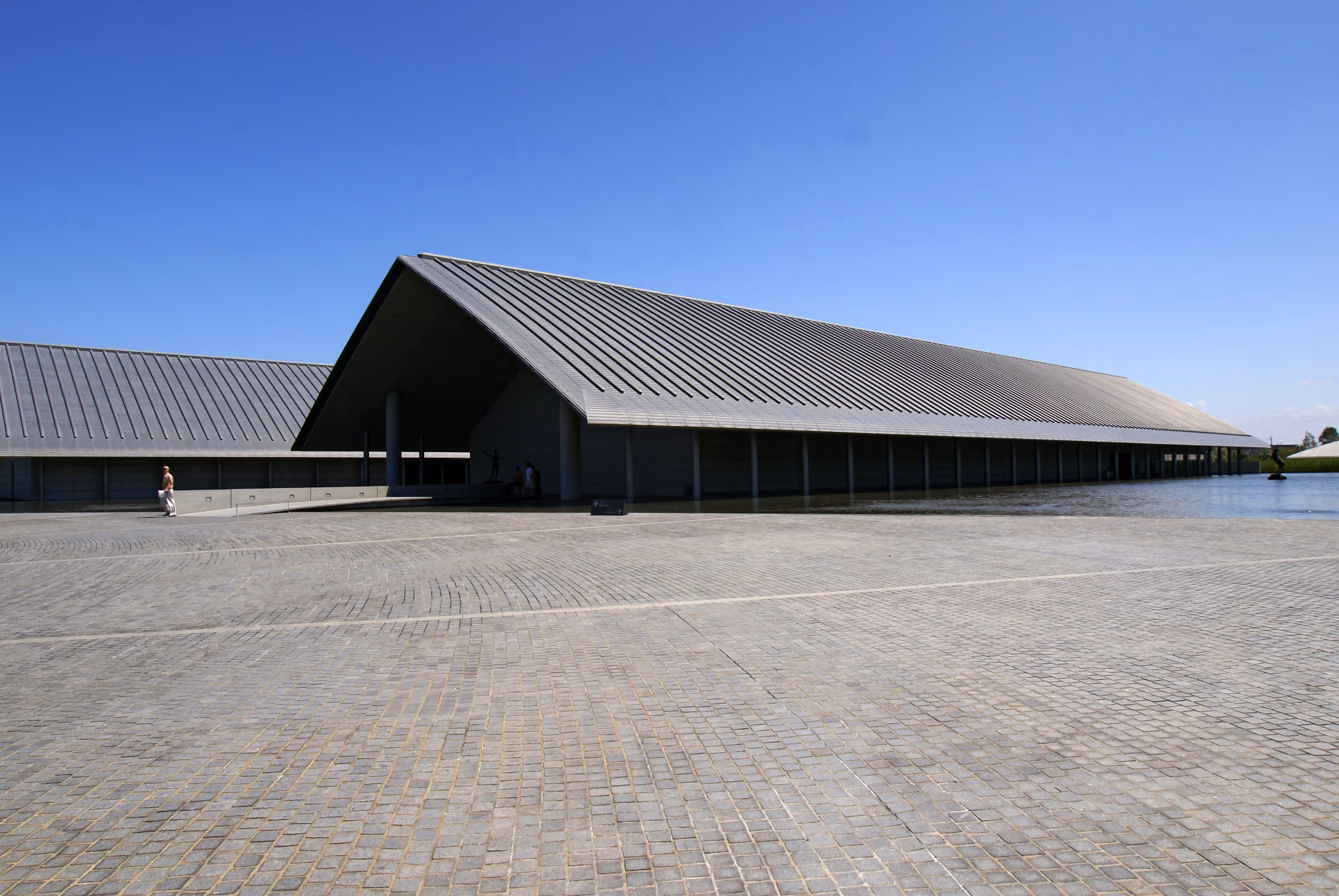 Modern building with a large sloped metal roof next to a reflecting pool under a clear blue sky.