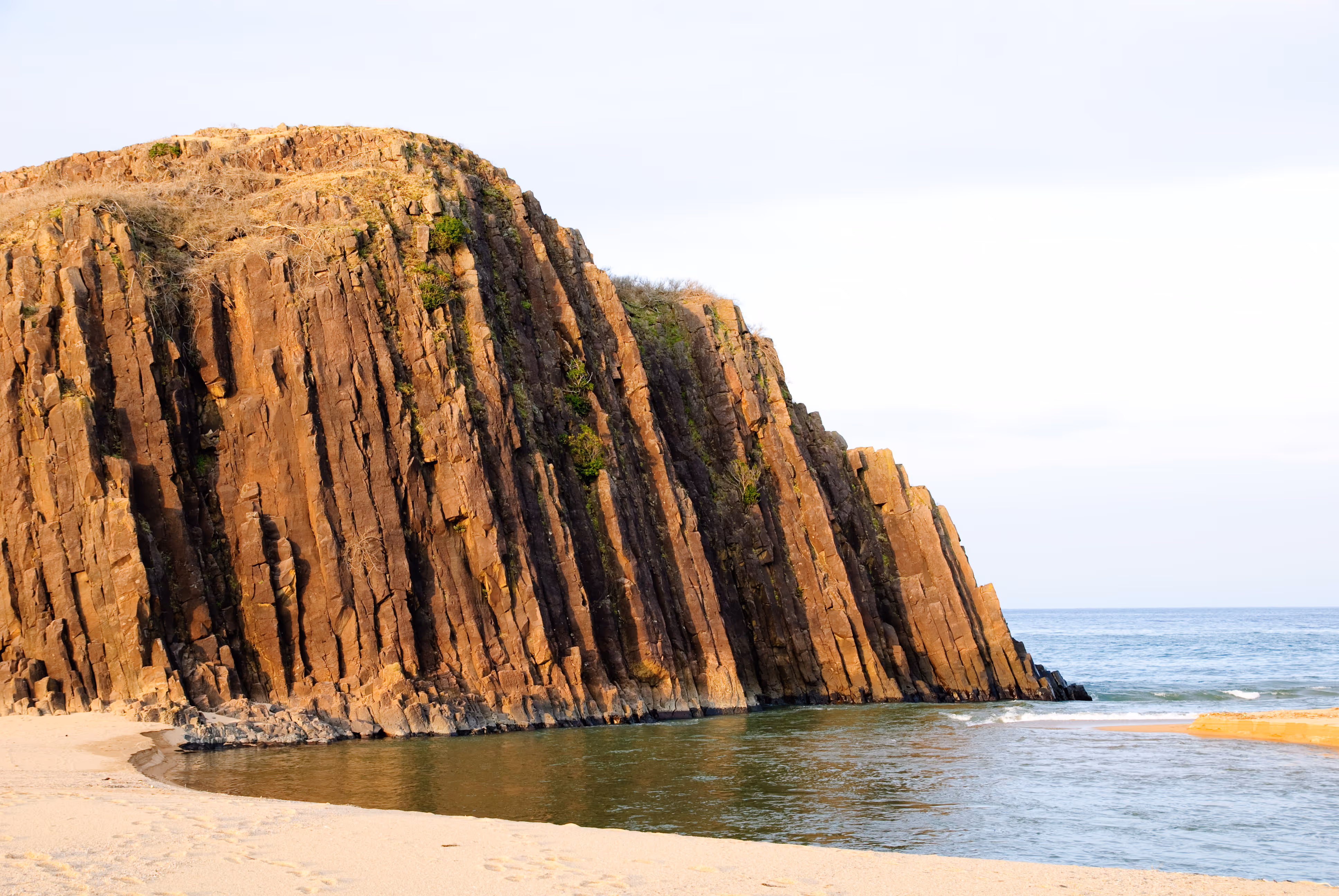 Rock formation with vertical columns by the ocean shore and sandy beach in the foreground.