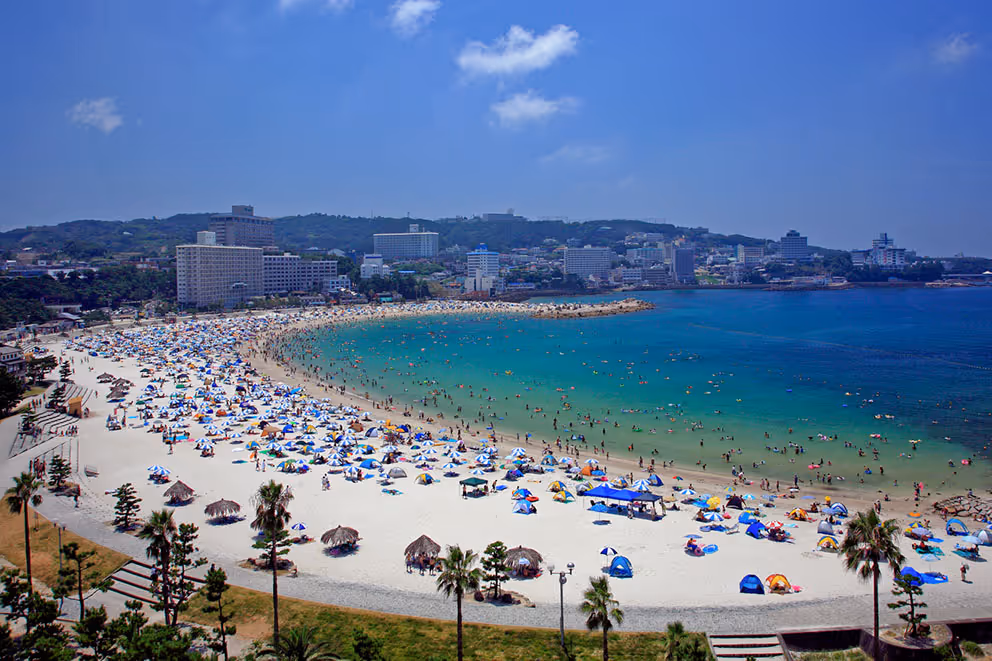 Crowded Shirahama Beach with white sand, blue umbrellas, people swimming in clear turquoise water, and a cityscape with hills in the background under a blue sky.