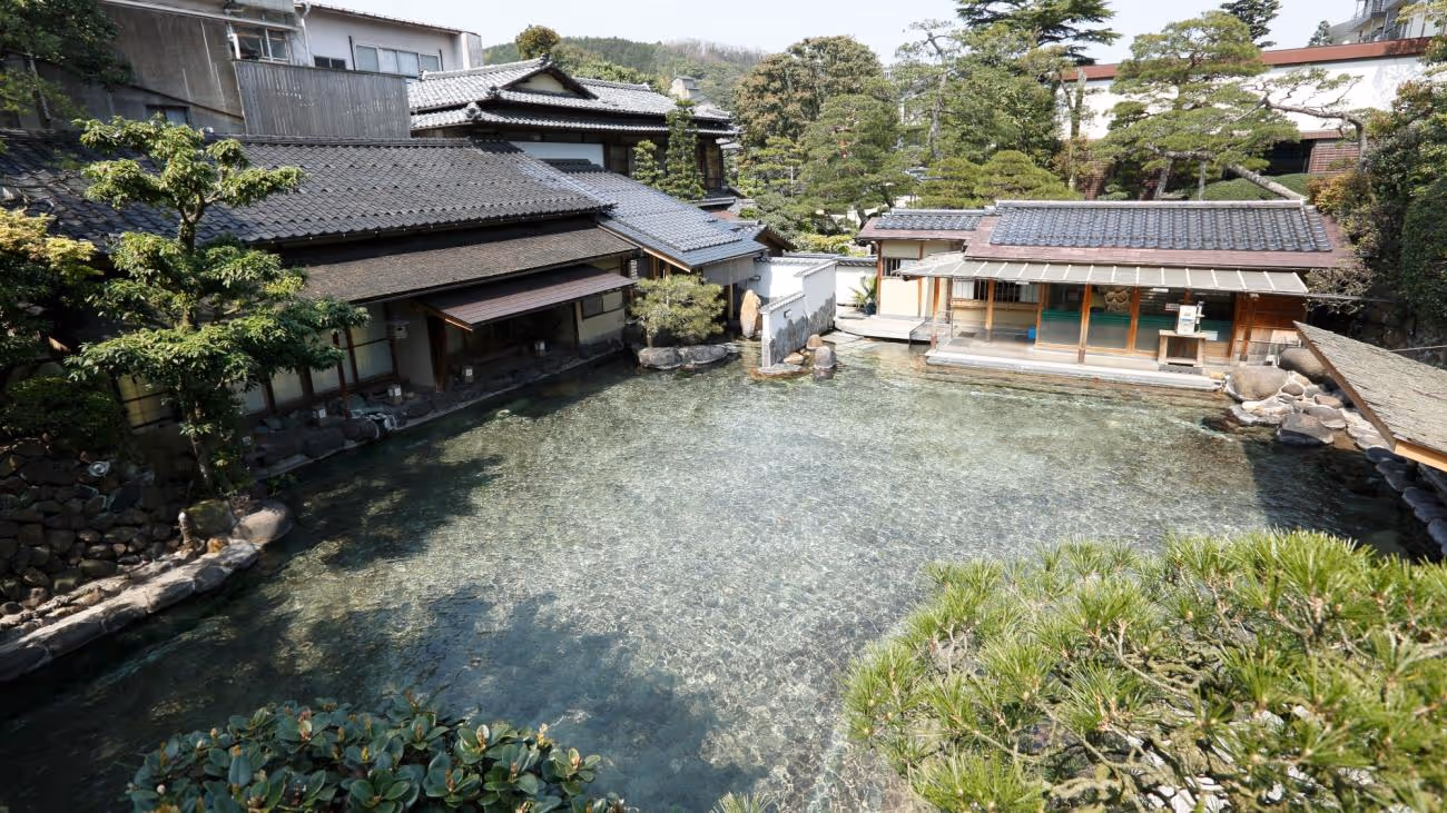 Traditional Japanese buildings surrounding a clear outdoor hot spring pool with green trees and shrubs.
