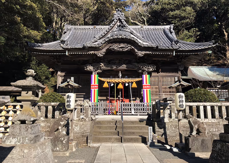 Front view of a traditional wooden Shinto shrine with ornate tiled roof, stone lanterns, and colorful banners in a forested area.