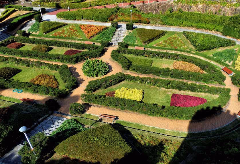 Aerial view of a manicured garden with geometric flower beds, hedges, walking paths, and benches under bright sunlight.