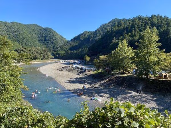 River flowing through a green forested valley with people swimming and camping along the rocky shore under a clear blue sky.