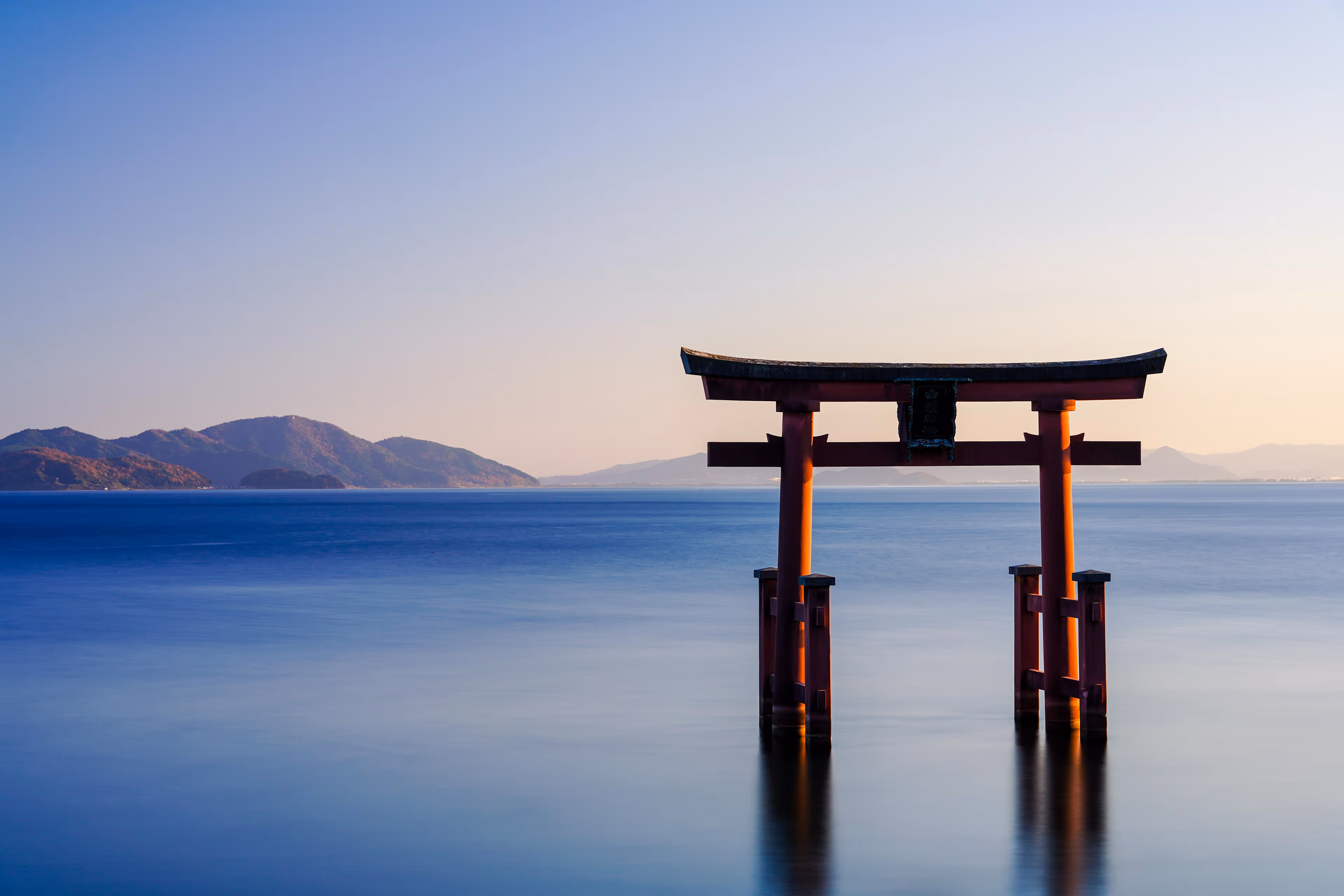 Red torii gate standing in calm blue water with distant mountains under a clear sky at sunset.