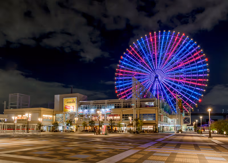 Colorfully lit Ferris wheel glowing in blue and red lights against a dark cloudy night sky over a spacious plaza with a shopping building.