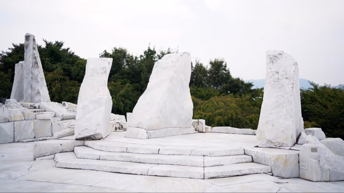 White marble stone monument on a stepped platform surrounded by green trees under a cloudy sky.