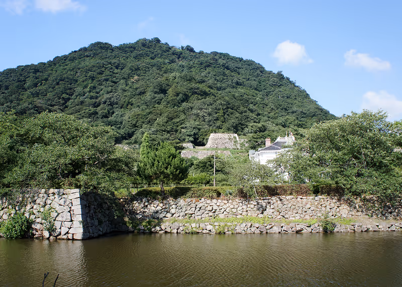 Stone ruins with green trees and a large forested hill in the background under a blue sky.