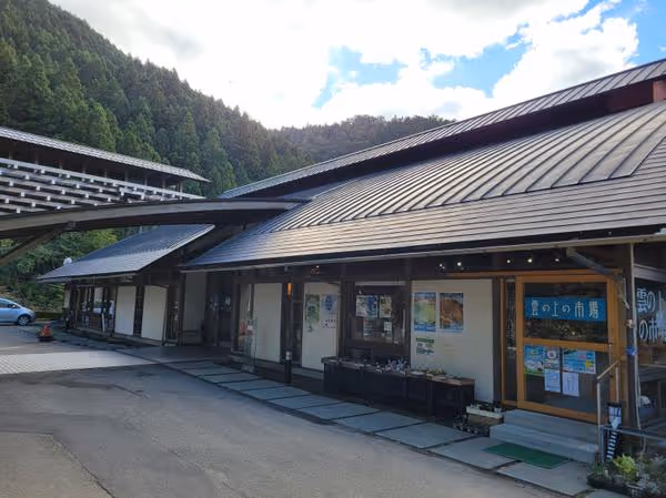 Traditional Japanese-style building with wooden exterior and metal roof set against green forested hills under a partly cloudy sky.